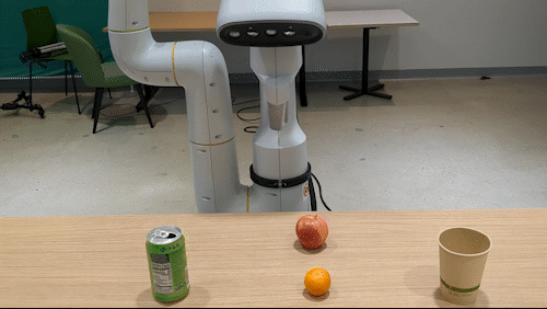 Robotic arm in a classroom setting with a can, apple, orange, and paper cup arranged on a table in front.